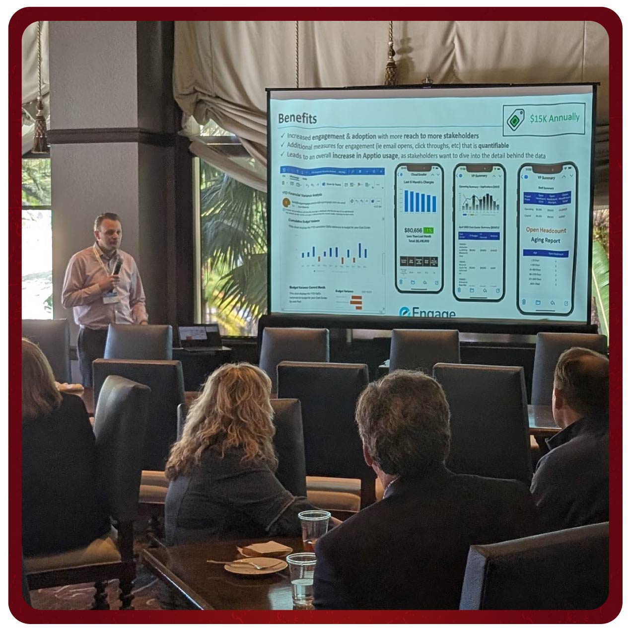 A speaker presents data on a large screen to attendees during a Rego University session. The screen displays graphs and metrics with relevant data. The attendees are seated around a conference table, engaging with the presentation.
