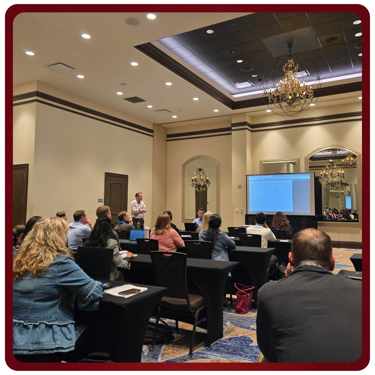 A speaker presents data on a large screen to attendees during a Rego University session. The attendees are seated around a conference table, engaging with the presentation.