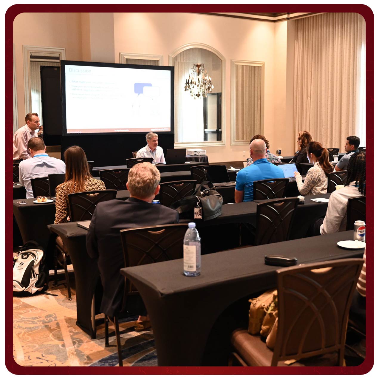 Two presenters listen to attendees sharing during a Rego University class. The attendees are seated at tables, engaging with the presentation.