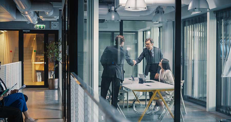 Business Leaders Complete a Job Interview With a Confident Hands Two colleagues shake hands during a meeting with a third colleague in a glass-walled office.